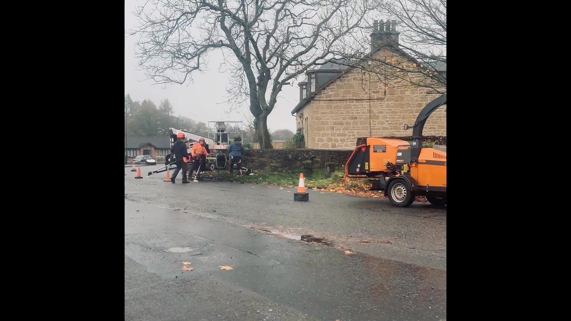 Gallery #9 Tree surgeons using a cherry picker platform and a wood chipper to work on a large bare tree beside a stone house.