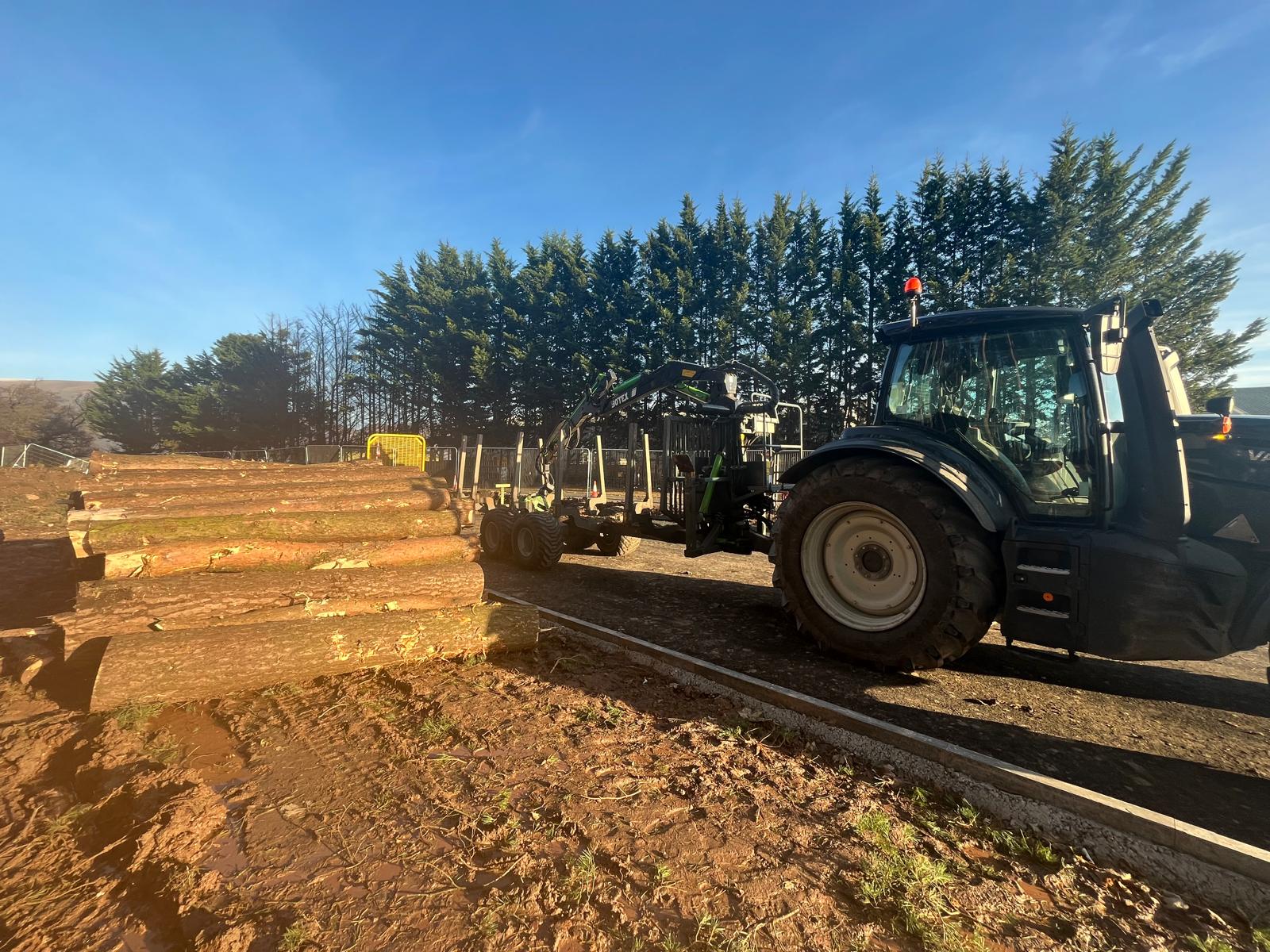 A tractor fitted with a forestry crane loads a stack of cut timber logs on site.