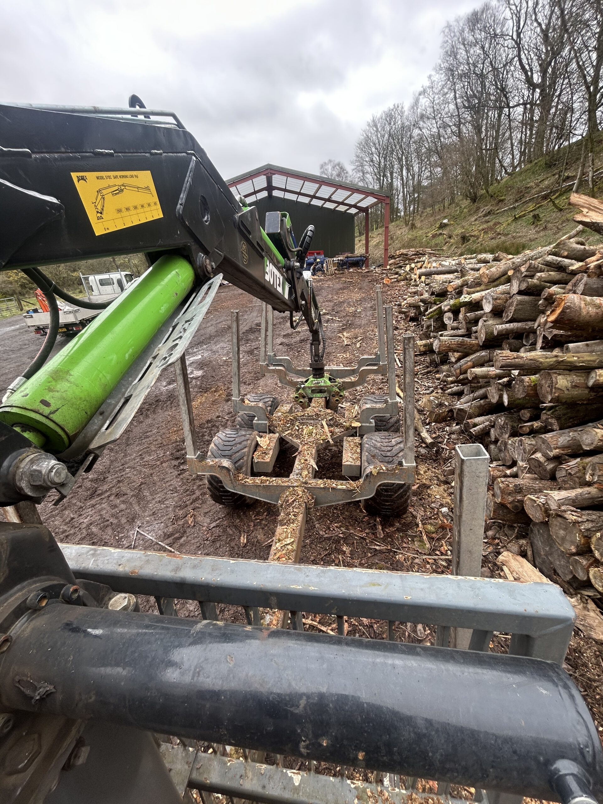 Hydraulic grapple loader stacking felled timber logs beside a storage shed at a forestry yard.