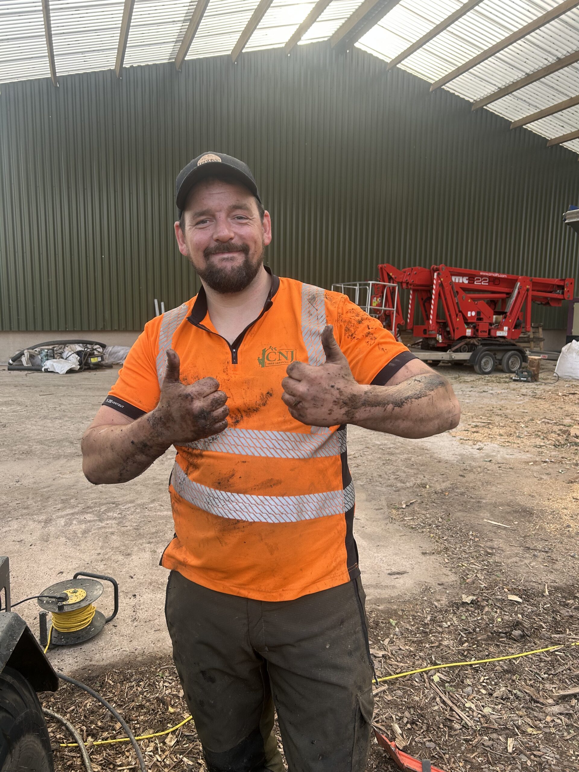 A muddy-handed CNJ Tree Services worker gives two thumbs up in front of yard equipment after a day&rsquo;s work.