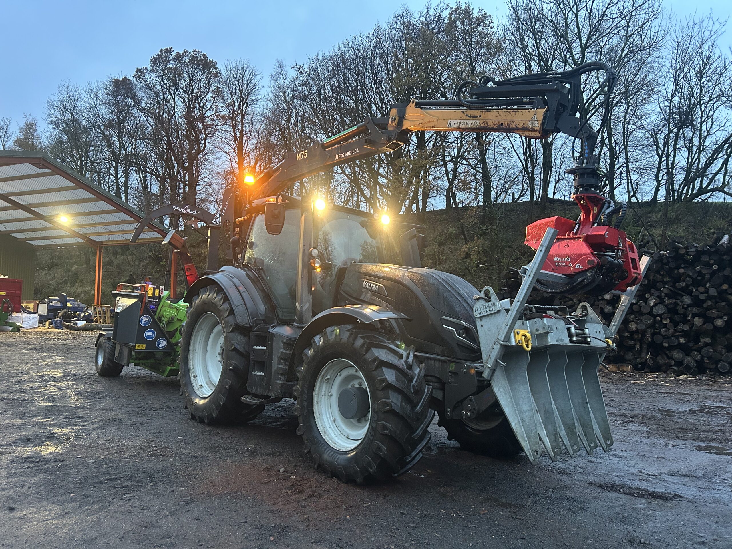 A Valtra tractor fitted with a timber grapple and wood chipper attachment stands in the yard at dusk beside stacked log piles.
