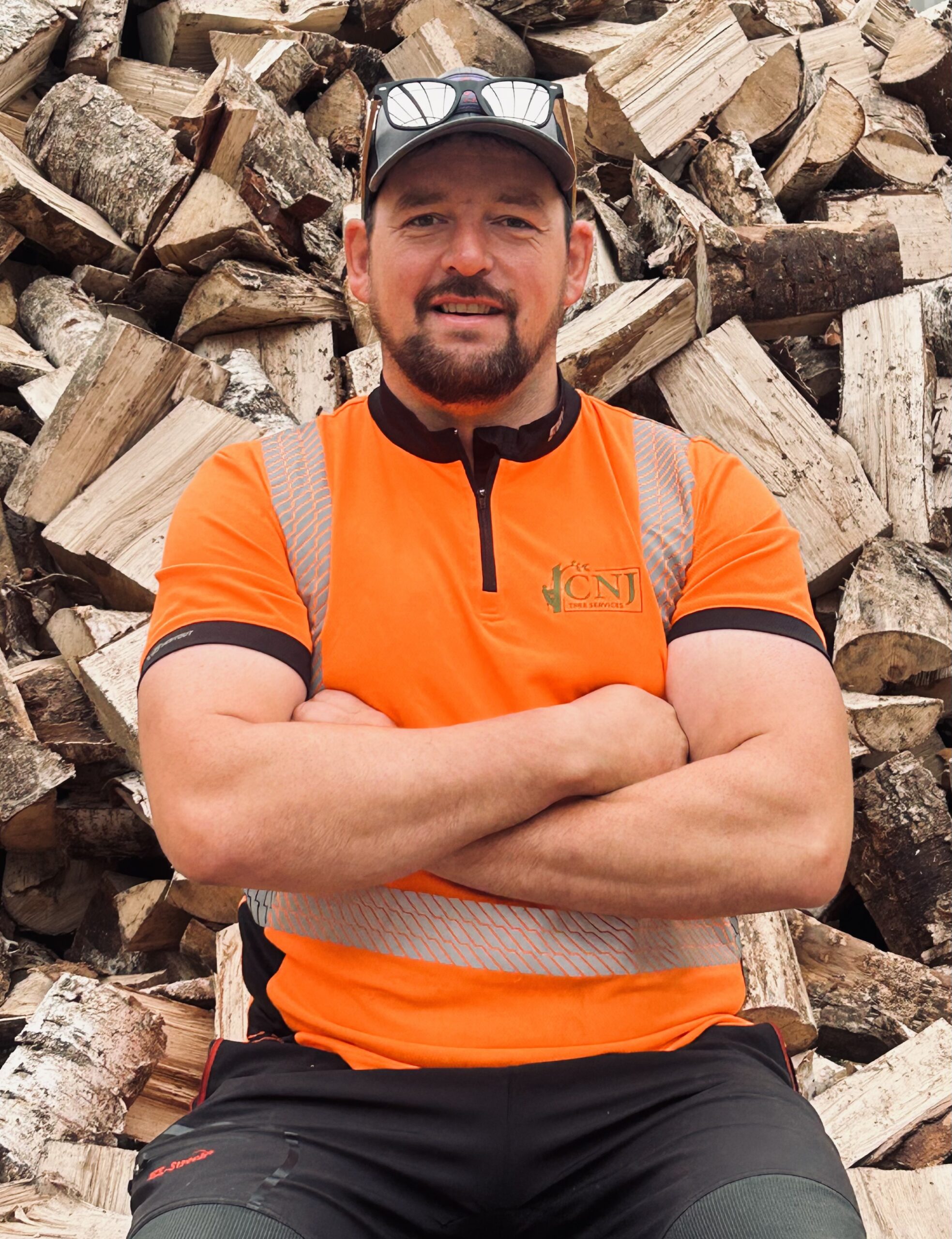 A CNJ Tree Services arborist stands with arms crossed in front of a large stack of cut firewood logs.