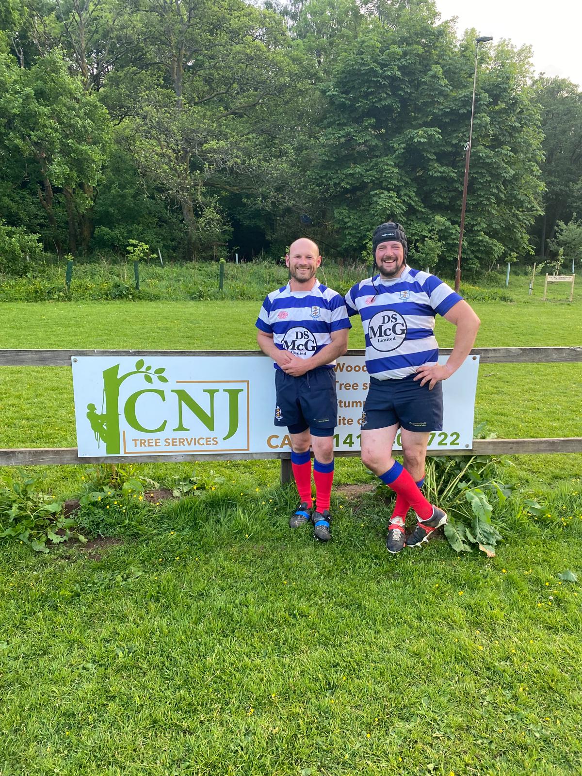 Two men in blue and white rugby kit pose beside a CNJ Tree Services sponsorship sign on a sports field.