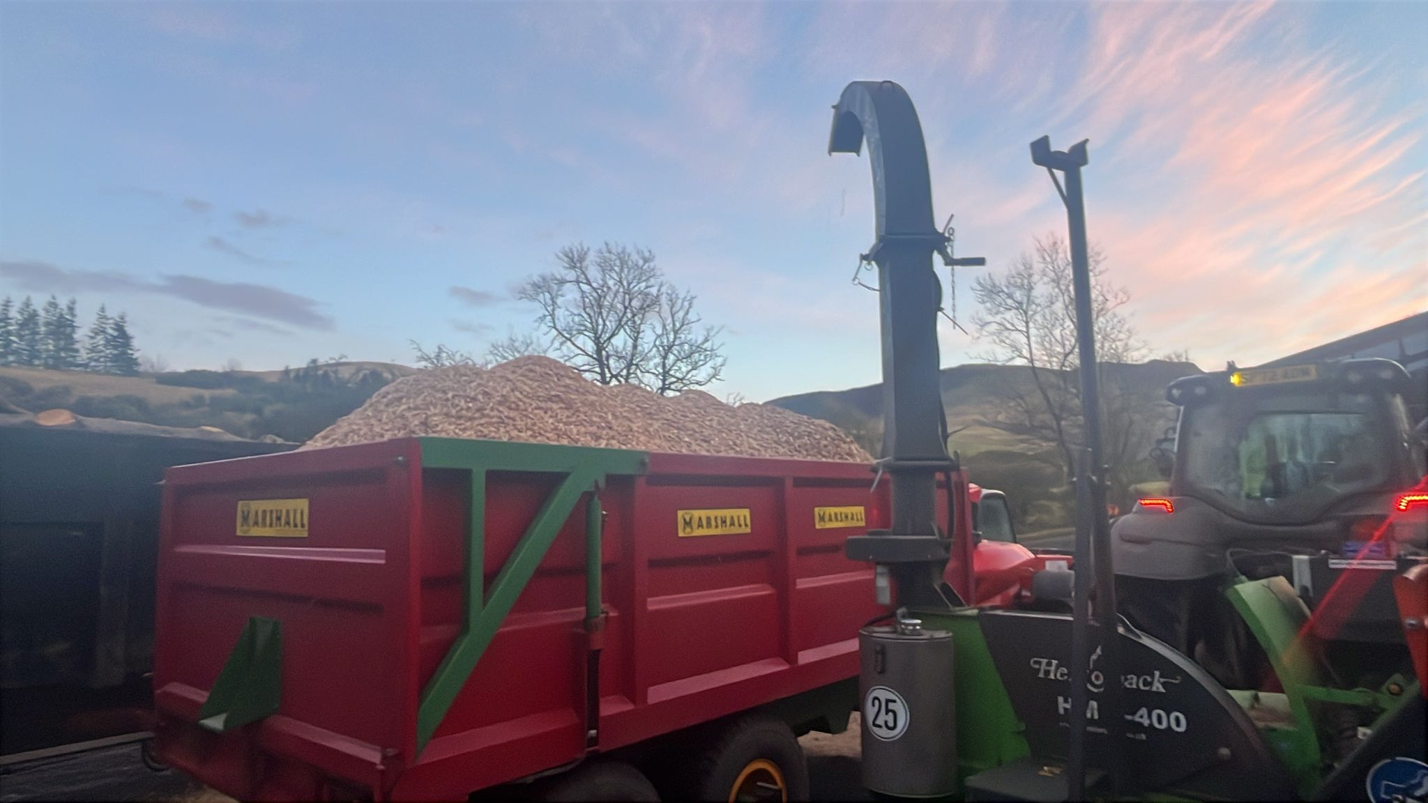 A large pile of wood chips is loaded into a red Marshall trailer using a chipper blower at dusk.