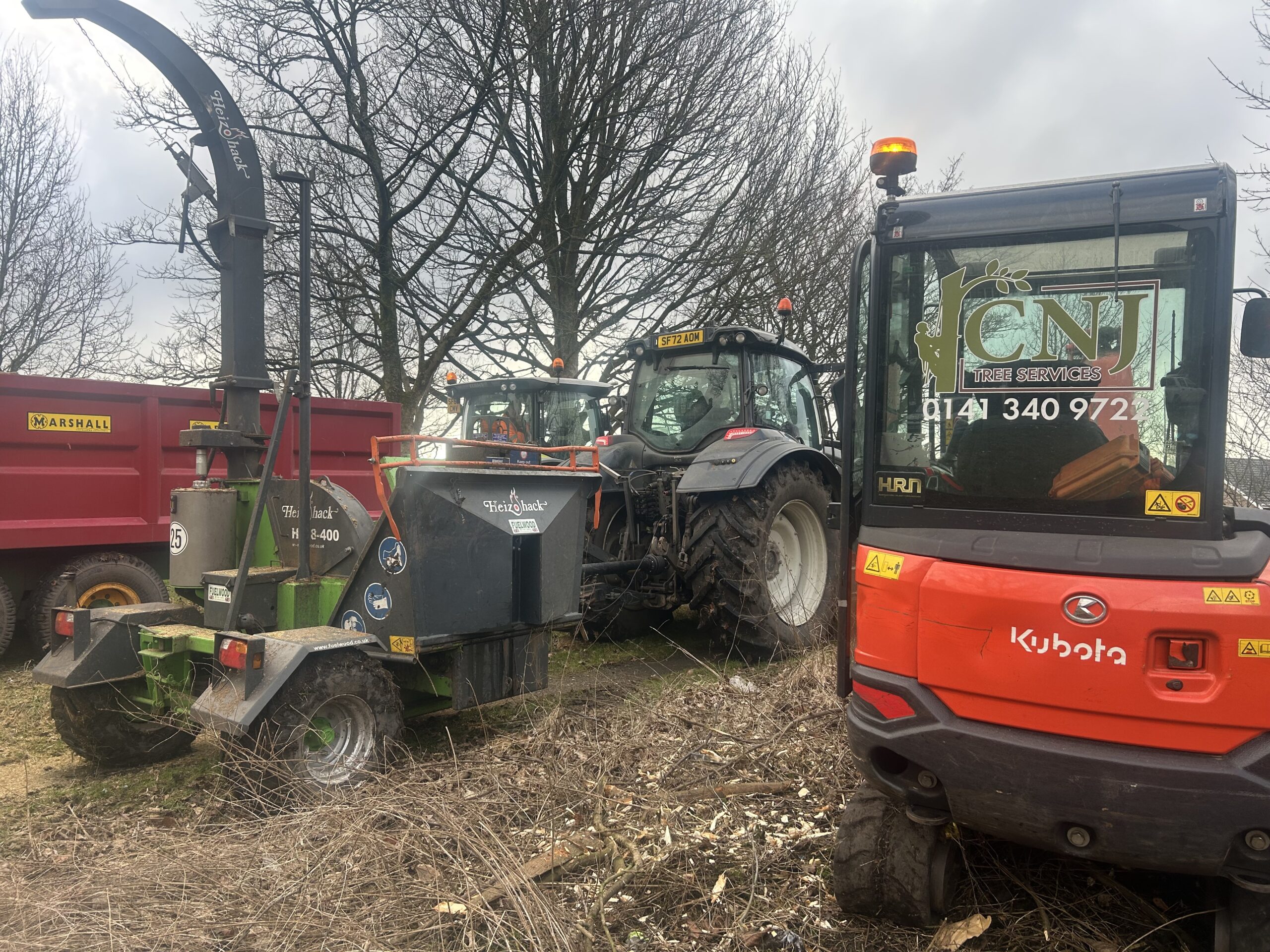 A wood chipper, tractors, and a CNJ Tree Services branded mini excavator parked together at a forestry work site.