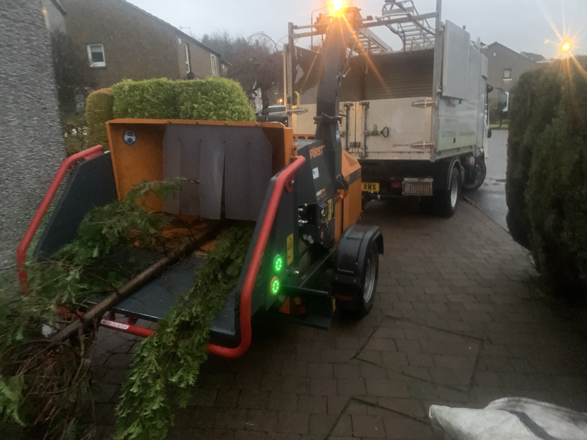 Gallery #54 An orange and black wood chipper loaded with cut conifer branches, parked in front of a tipper truck at dusk.