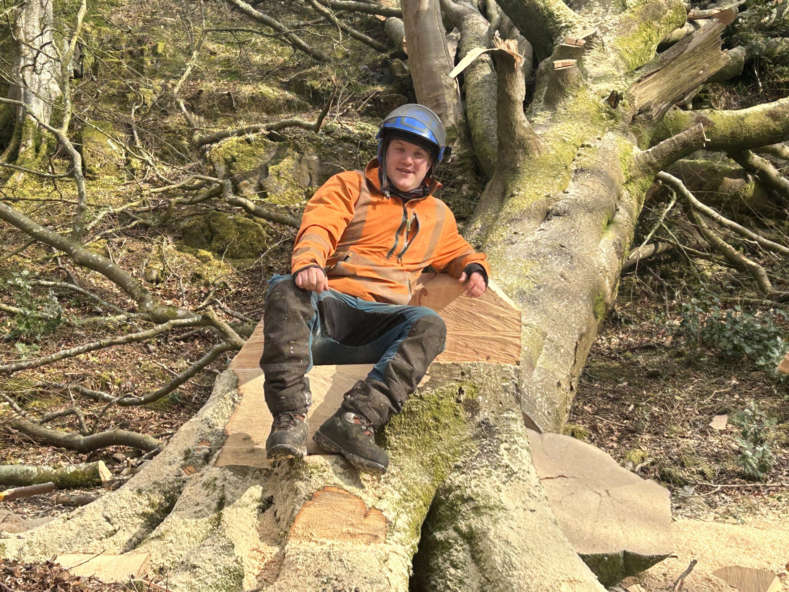 Gallery #55 A smiling tree surgeon in helmet and high-vis jacket sits on a large felled tree trunk in the woods.